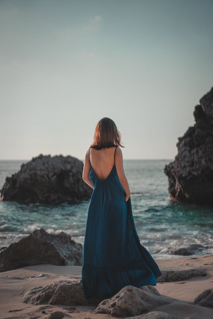 Back view full body fashionable female wearing blue maxi dress standing on rocky seacoast against azure rippling sea on clear summer weather